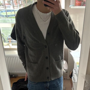 Grå/grön kofta med knappar - Mysig grå kofta med v-ringning och stora svarta knappar framtill. Koftan har två fickor och ribbade muddar vid ärmslut och nederkant. Perfekt att slänga på sig över en t-shirt för en chill och stilren look. Priset är diskuterbart