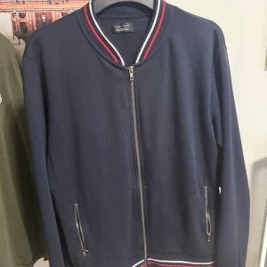 Zip kofta från Zara Man - Snygg mörkblå tröja från Zara Man med dragkedja framtill och två fickor med dragkedjor. Krage och nederkant har randiga detaljer i vitt och rött. Koftan har lång ärm och en clean look. 