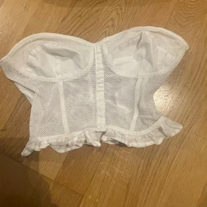 Vit korsett från Guess med volang - Säljer en vit korsett från Guess med meshdetaljer och snygg volangkant nedtill. Toppen är axelbandslös och har en strukturerad form med markerade kupor. Perfekt för dig som vill ha en trendig och feminin look.