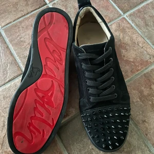 Christian Louboutin svarta sneakers med nitar - Svarta sneakers från Christian Louboutin i mocka med coola svarta nitar på tån och klassisk röd sula med logotyp. Skorna har snörning och rund tå, vilket ger en edgy vibe. Perfekta för dig som vill sticka ut med exklusiv streetstyle.