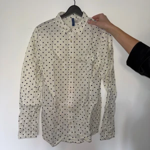 Vit prickig skjorta från H&M Divided - Snygg vit skjorta med svarta prickar från H&M Divided. Klassisk krage, långärmad och knäppning framtill samt vid ärmslut. Tillverkad i mjuk bomull som känns skön mot huden. Perfekt för dig som gillar coola mönster och vill sticka ut lite extra.