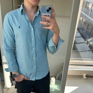 Ljusblå Ralph Lauren skjorta  - Snygg ljusblå skjorta från Ralph Lauren i slim fit med klassisk krage och knappar framtill. Skjortan har lång ärm och ikoniska Polo-loggan broderad på bröstet. Perfekt för dig som gillar stilrena och tidlösa plagg. Materialet är mjuk bomull.