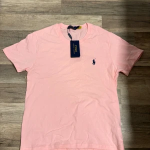 Rosa t-shirt från Polo Ralph Lauren - Snygg rosa t-shirt från Polo Ralph Lauren med klassisk rund hals och broderad marinblå logga på bröstet. Tillverkad i mjuk bomull och har en normal passform. Perfekt för en clean och stilren look.