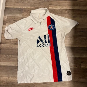 PSG vit bortatröja Nike Man  - Paris Saint-Germain bortatröja i vitt från Nike med klassisk krage, röda och blå vertikala ränder samt klubbmärke och sponsortryck på bröstet. Tillverkad i lätt och ventilerande polyester med Nike Vaporknit-teknologi. Perfekt för dig som älskar fotboll och PSG.