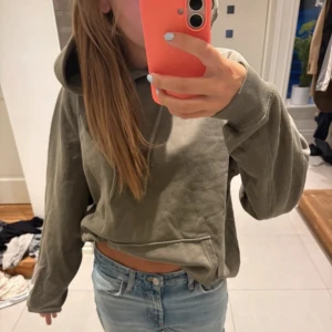 Olivgrön croppad hoodie - Säljer en olivgrön croppad hoodie med huva och magficka. Tröjan har en loose passform och långa ärmar, perfekt för chill dagar. Materialet är mjukt och skönt, troligen bomull eller polyester. Passar dig som gillar avslappnad stil. avklippt längst ner men inget man tänker på