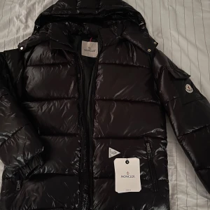 Moncler maya - (HERR JACKA)Snygg svart pufferjacka från Moncler med glansig finish, huva och dragkedja framtill. Jackan har klassisk Moncler-logga på ärmen och coolt serietryck på insidan. Perfekt för kalla dagar och riktigt stilren look.