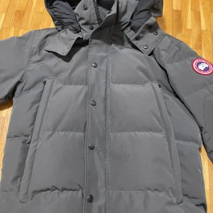 Mörkgrå Canada Goose jacka - Säljer en mörkgrå jacka från Canada Goose med huva och klassisk logopatch på ärmen. Jackan har quiltad design, dragkedja och knappar framtill samt två stora fickor. Perfekt för kalla vinterdagar och riktigt snygg till streetwear. Ett problem är att dom 2 knapparna längst ner har gått sönder och har inte päls därför släjs den billigt