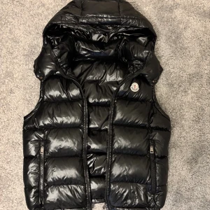 Svart dunväst från Moncler - Snygg svart dunväst från Moncler med glansig finish och huva. Västen har två dragkedjefickor framtill och Moncler-logga på bröstet. Perfekt för lager-på-lager och håller dig varm under kyliga dagar. Stängs med dragkedja framtill.