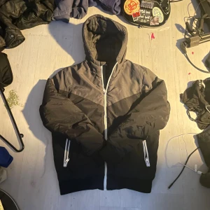 Svart och grå pufferjacka från FSBN - Säljer en stilren pufferjacka från FSBN i svart och grått. Jackan har huva, vita dragkedjor och coola detaljer på ärmen. Perfekt för dig som vill ha en varm och snygg jacka med modern look. Passar dig som gillar streetwear och enkel design.