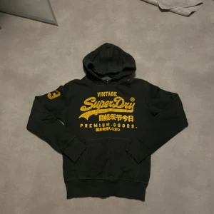 Grön Hoodie Superdry med gul print - Svart hoodie från Superdry med stor gul vintage-print på bröstet och japanska tecken. Siffran 23 i gult på vänster ärm. Klassisk huva och känguruficka framtill. Mjuk bomullskvalitet och ribbade muddar vid ärmslut och nederkant.