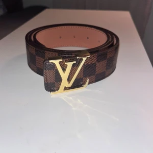 Louis Vuitton bälte - Säljer ett snyggt Louis Vuitton bälte i brunt och svart med klassiskt rutigt mönster. Bältet har en ikonisk LV-spänne i guld och är tillverkat i skinn. Perfekt accessoar för att lyfta din outfit med lyxig känsla och stil. Använd 4ggr.