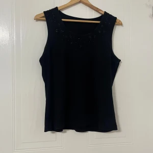 Svart ärmlös topp med pärlor - Säljer en svart ärmlös topp med rund hals och dekorativa svarta pärlor och små paljetter längs halsringningen. Toppen har en klassisk, rak passform och är gjord i mjukt material som känns skönt mot huden. Perfekt för dig som gillar stilrena detaljer.