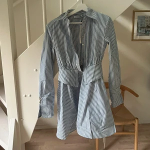 Blåvit randig skjortklänning Adoore - Snygg blåvit randig skjortklänning från Adoore med markerad midja och långa ärmar. Klänningen har klassisk krage, knappar framtill och breda manschetter. Perfekt för dig som gillar en stilren och trendig look.