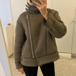 Brun bikerjacka från Zara med dragkedjor - Säljer en brun bikerjacka från Zara med oversized passform, stora silvriga dragkedjor och mysigt teddyfoder. Jackan har hög krage med spännen, detaljerade sömmar och justerbara spännen i sidorna. Perfekt för kalla dagar!