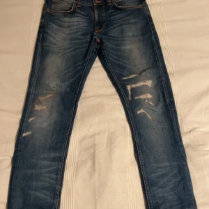 Nudie Jeans med slitningar - Säljer ett par blå jeans från Nudie Jeans Co i storlek W31 L32. Jeansen har slitna detaljer framtill, klassisk femficksmodell och orangea sömmar. De är tillverkade i denim och har en rak passform med snyggt tvättad look. Modellen är lean Dean och är i nyskick, det finns inga hål utan bara ytliga slitngar och as skön wash o fade.