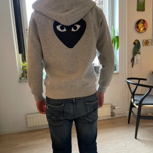 Grå hoodie Comme des Garçons Play - Säljer en grå hoodie från Comme des Garçons Play med dragkedja och huva. Ikoniskt svart hjärta med ögon på bröstet och stort tryck på ryggen. Mjuk insida och kängurufickor framtill. Perfekt för chill dagar och streetstyle.