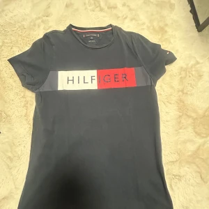Blå Tommy Hilfiger t-shirt - Snygg mörkblå t-shirt från Tommy Hilfiger med klassiskt logotryck i vitt, rött och blått över bröstet. T-shirten har rund halsringning och korta ärmar. Perfekt för en stilren och avslappnad look.