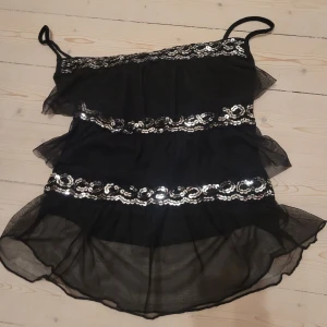 Svart paljett-topp med volanger - Supersöt svart topp  med tunna axelband och flera lager volanger. Toppen har glittriga paljettband i silver som ger en extra festlig känsla. Tunt, transparent tyg som faller snyggt och ger en luftig look.