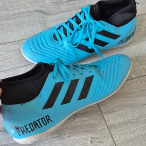 Adidas Predator blå fotbollsskor - Säljer ett par Adidas Predator fotbollsskor i en snygg blå färg med svarta detaljer och vita sulor. Skorna har snörning, klassiska tre ränder på sidan och texten 'Predator' på hälen. Ovandelen är i syntetmaterial och insidan har en svart, stretchig krage för extra komfort.