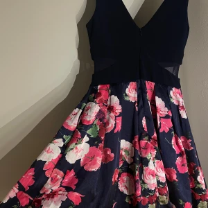 Marinblå blommig kortklänning - Supersnygg marinblå kortklänning med v-ringad överdel och mesh-detaljer i midjan. Kjolen är utsvängd med stora rosa och vita blommor på mörkblå botten. Materialet känns glansigt och har en lyxig känsla, perfekt för dig som vill sticka ut med färg och mönster.