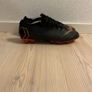 Nike Mercurial svarta fotbollsskor - Svarta Nike Mercurial fotbollsskor med orange detaljer och logga. Skorna har snörning, platt sula med dobbar och är tillverkade i syntetmaterial. Perfekta för dig som vill ha snabba och lätta skor på planen. Skicka även frågor samt prisförslag 