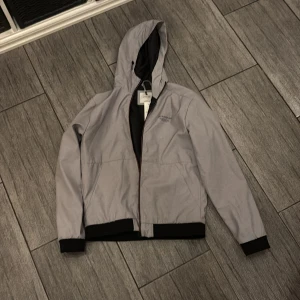 Ljusgrå vindjacka från Jack & Jones - Ljusgrå vindjacka med huva från Jack & Jones. Aldrig använd, helt ny. Originalpris är 700kr och prislappen finns fortfarande kvar. Hör gärna av dig om du är intresserad. Mvh Gustav