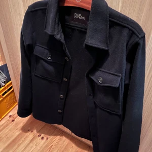 Svart overshirt från Oscar Jacobson - Stilren svart overshirt från Oscar Jacobson med klassisk krage, två bröstfickor med lock och knappar framtill. Jackan är tillverkad i mjuk ull och har en rak passform som funkar till många olika looks. Har en liten skavank på vänster underarm men märks inte om man inte kollar noga