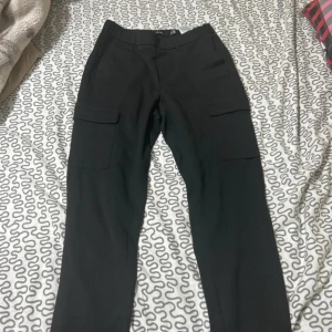 Svarta cargopants med fickor från Gina Tricot - Svarta cargopants med raka ben och stora sidofickor. Byxorna har en klassisk midja och är tillverkade i ett mjukt bomullsmaterial. Perfekta för en chill och avslappnad stil, med praktiska fickor för mobilen och annat.