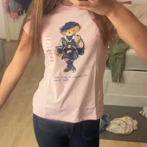 Rosa Polo Bear t-shirt från Ralph Lauren - Söt ljusrosa t-shirt från Ralph Lauren med ikoniska Polo Bear-trycket framtill. Klassisk passform och korta ärmar, perfekt till jeans eller kjol. Mjuk bomull som känns skön mot huden. En riktig statement-piece för dig som gillar gulliga och trendiga plagg. Kun brukt noen ganger