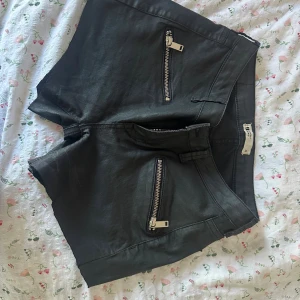 Svarta shorts med dragkedjor från g perfect jeans - Snygga svarta shorts från g perfect jeans i storlek 38. De har två coola dragkedjor framtill och en lite glansig finish som ger en edgy vibe. Perfekta för dig som gillar en tuffare stil och vill sticka ut lite extra.