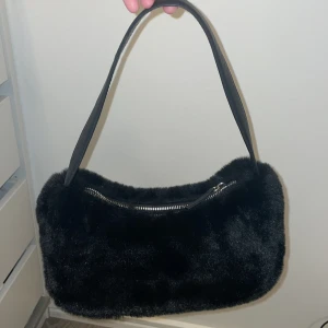 Svart pälsig axelväska från H&M - Snygg svart axelväska från H&M med mjuk pälsig yta och silverfärgad dragkedja. Väskan har en enkel, avlång form och ett stilrent handtag i svart. Perfekt för dig som vill ha en trendig accessoar med lite extra attityd.