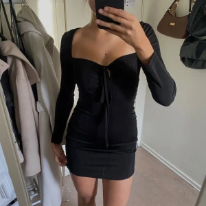 Svart tight miniklänning - Supersnygg svart miniklänning med lång ärm och tight passform. Klänningen har en fyrkantig urringning och snörning framtill. Klänningen är lite glittrig. Inga defekter