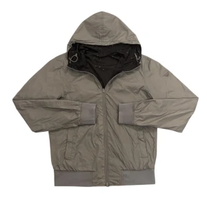 Armani Windbreaker  - Snygg vändbar vindjacka från Armani Jeans med huva. Ena sidan är grå med diskret logga på ärmen, andra sidan är svart med små mönstrade loggor. Jackan har dragkedja fram, två fickor med dragkedja och ribbade muddar. Perfekt för dig som vill ha en clean och stilren look.