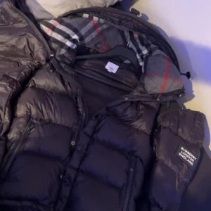 Svart pufferjacka från Burberry S - Säljer en svart pufferjacka från Burberry med klassiskt rutigt foder i huvan. Jackan har dragkedja, tryckknappar och två snedställda fickor framtill. Materialet är nylon och polyester, perfekt för kalla dagar. Stilsäker patch med Burberry London England på ärmen.