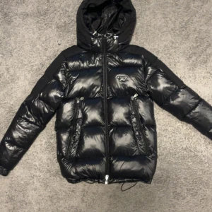 Svart pufferjacka Alessandro Zavetti - Svart pufferjacka från Alessandro Zavetti med glansig finish och huva. Jackan har två dragkedjeförsedda fickor fram, justerbar nederkant med snörning och mönstrat innerfoder med logotyp. Materialet är polyester och polyamid, perfekt för kalla dagar.