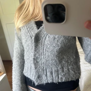 Grå stickad kofta med knappar - Mysig grå stickad kofta med tre stora knappar framtill. Koftan är croppad med bred ribbad kant nedtill och vid ärmslut. Koftan är i storlek Xs