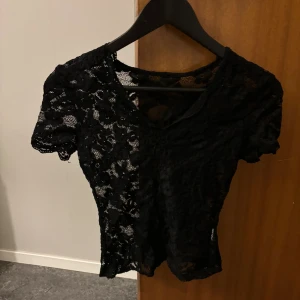 Svart spetsblus med kort ärm - Säljer en svart blus i genombruten spets med korta ärmar och v-ringning. Toppen har ett blommigt spetsmönster och är figurnära i modellen. Perfekt för dig som vill ha en snygg och stilren topp med lite edge.