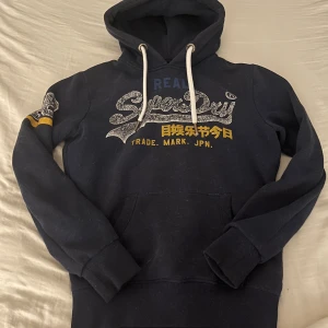 Mörkblå Superdry hoodie med tryck - Snygg mörkblå hoodie från Superdry med stort glittrigt tryck på bröstet och japanska tecken i gult. Tröjan har huva med vita snören, känguruficka och ribbade muddar. Perfekt för dig som gillar streetwear och vill ha en cool look.
