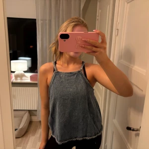 Blå denim linnetopp med smala axelband - Säljer en chill blå linnetopp i denim-look med smala axelband och avslappnad passform. Toppen har en enkel design med ljusare sömmar längs kanterna och är perfekt för varma dagar. Passar dig som gillar en laidback stil.