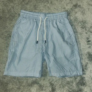 Blåvitrandiga shorts med snörning - Snygga blå shorts med vita vertikala ränder och elastisk midja med vit snörning. Lätta och luftiga, perfekta för varma dagar. Har sidofickor och en bakficka. Klassisk och fräsch look som passar till det mesta.