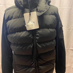 Svart dunväst från Moncler med huva - Svart dunväst från Moncler med quiltad framsida, huva och dragkedja. Västen har en snygg Moncler-logga på ärmen och praktisk ficka med knapp. Materialet är polyester och dun, perfekt för kyliga dagar. Stilsäker och sportig look med klassiska detaljer.