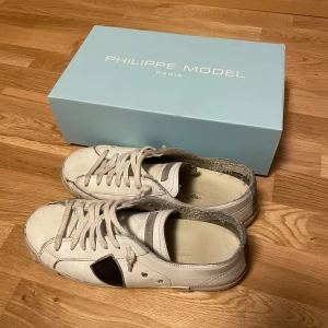 Vita Philippe Model sneakers med svart detalj - Säljer ett par vita Philippe Model sneakers i skinn med svart detalj på sidan och snörning framtill. Sulan har tydlig Philippe Model-logga undertill. Klassisk och clean design som passar till det mesta. Perfekta för dig som gillar stilrena och trendiga sneakers.