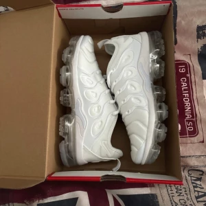 Nike Air Vapormax Plus vita sneakers - Nike Air Vapormax Plus i helvitt med futuristisk design och genomskinlig sula. Skorna har snörning, rund tå och är tillverkade i syntetmaterial med coola detaljer på ovandelen. Perfekta för dig som vill sticka ut med en modern sneaker-look.