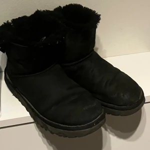 Svarta äkta UGG boots i fårskinn - Mysiga svarta UGG boots med fluffigt foder och rund tå. Skorna är tillverkade i mjukt fårskinn och mocka, med en platt sula som ger skön komfort. Perfekta för kalla dagar när du vill hålla fötterna varma och stilrena.