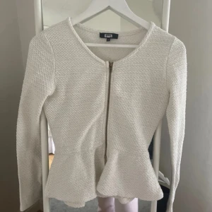 Stickad kofta  - Säljer en vit kofta från bik bok med snygg struktur, rund halsringning och guldfärgad dragkedja framtill. Modellen har peplumvolang nedtill och långa ärmar. Perfekt för dig som gillar stilrena och feminina detaljer.xs/s