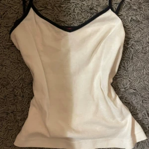 Vit ribbad linnetopp med svarta kanter - Säljer en vit ribbad linnetopp med svarta kanter och smala axelband. Toppen har en v-ringad hals och ärmlös design, perfekt för att styla med jeans eller kjol. Materialet känns mjukt och följsamt mot huden.