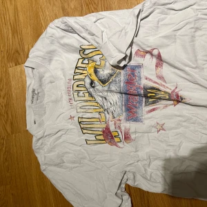 Vit Wrangler t-shirt med örntryck - Vit t-shirt från Wrangler med stort vintageinspirerat tryck framtill. Motivet har en örn, texten 'Wilderness' och färger i gult, rött och blått. Klassisk rund hals och korta ärmar. Perfekt för dig som gillar retrostil och statement-plagg.