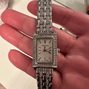 Silverfärgad  klocka  - Snygg rektangulär klocka i silverfärg med glittrande stenar runt urtavlan och på länken. Urtavlan har ett diskret rutmönster och minimalistiska markeringar. Perfekt accessoar för att lyfta din stil.