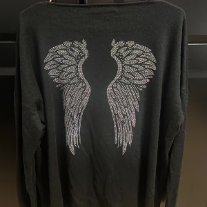 Svart stickad tröja med vingar AVA - Svart stickad tröja från AVA by Paul Stragas med v-ringning och långa ärmar. Baksidan har snygga silvriga änglavingar i paljettdetaljer. Ett litet, nästan osynligt hål på framsidan. Lappen i nacken saknar storlek, men jag uppskattar att den motsvarar ungefär storlek L-XL. Perfekt för vardagsbruk!