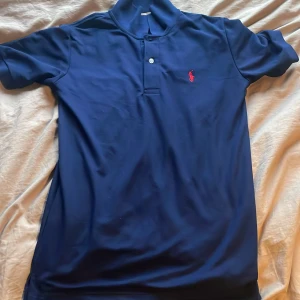 Marinblå pikétröja från Ralph Lauren - Snygg marinblå pikétröja från Ralph Lauren med klassisk krage och två knappar framtill. Tröjan har det ikoniska röda broderade polospelar-loggan på bröstet. Perfekt för en clean och sportig stil.